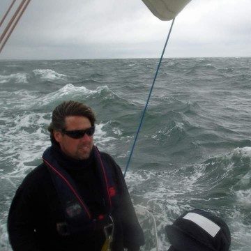 Ein Mann mit Sonnenbrille steht auf einem Segelboot, umgeben von stürmischer See.