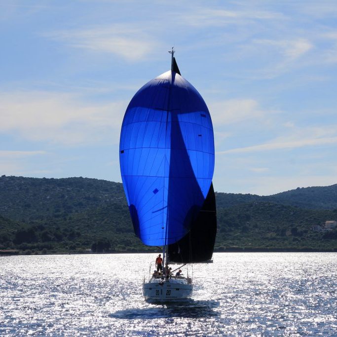 Segelboot mit blauen Segeln auf glitzerndem Wasser, im Hintergrund Hügel.